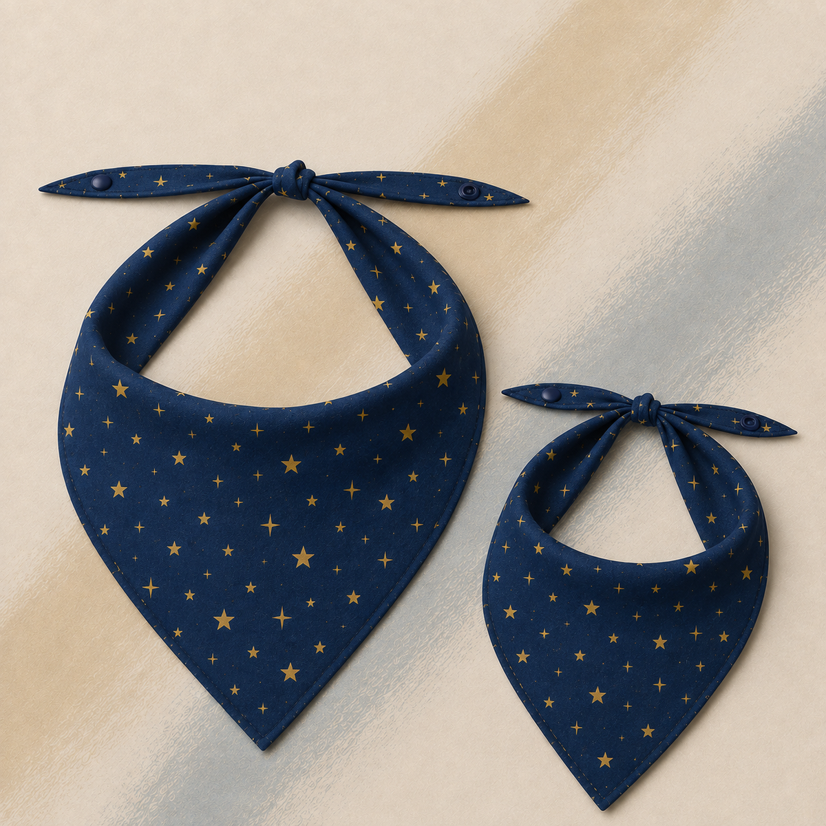 Two navy blue fabric bibs with a gold star pattern, one large and one small, displayed on a textured beige background.