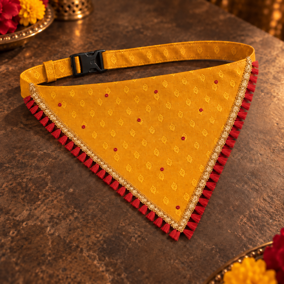 Mustard yellow and red ethnic dog bandana with woven gold trim and an adjustable buckle, displayed flat on a brown stone surface.

